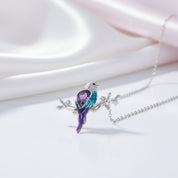 A silver bird pendant necklace perched on a small branch, featuring a purple crystal body and blue-green crystal wings, displayed on a soft white and pale pink satin background.