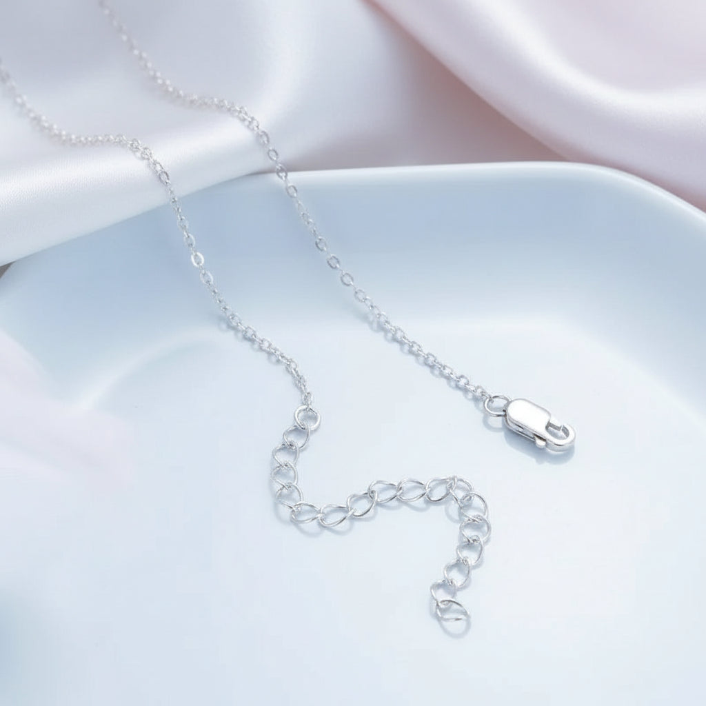 A delicate silver chain necklace with a lobster clasp, displayed on a light blue dish with soft pink satin fabric in the background.