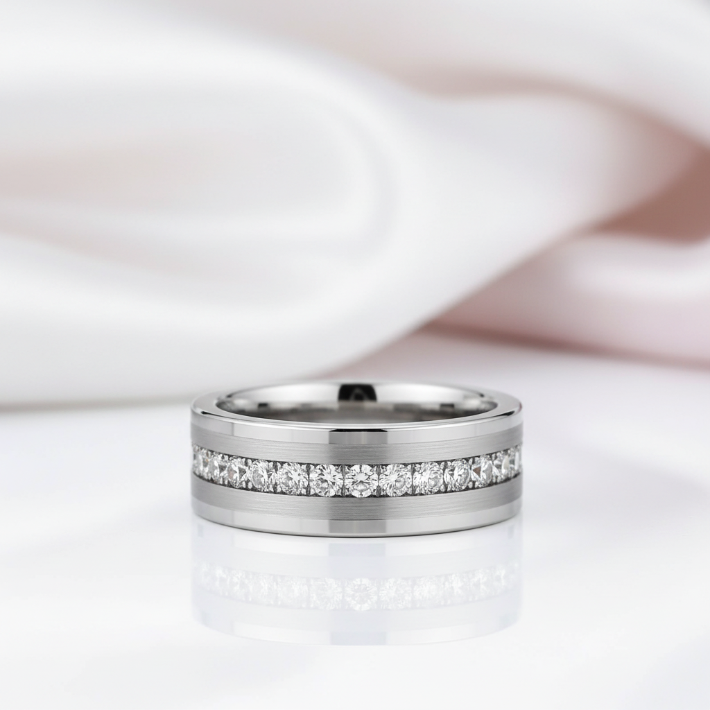 A polished silver-toned band ring with a channel-set row of clear stones, photographed on a white reflective surface with soft pink fabric in the background.