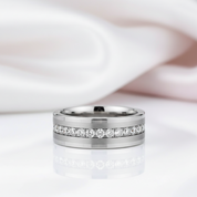 A polished silver-toned band ring with a channel-set row of clear stones, photographed on a white reflective surface with soft pink fabric in the background.