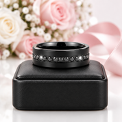 A black metal band ring with a row of small dark gemstones, displayed on a black jewelry box with soft pink roses and a pink ribbon in the background.
