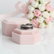 A silver-toned ring with a dark floral inlay sits on a small pink jewelry box, with another pink box and a bouquet of pink and white roses blurred in the background.