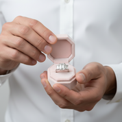 A close-up of a person in a white shirt holding an open pale pink octagonal ring box that displays a silver-toned ring with three clear rectangular gemstones.