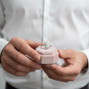 Person holding a pink jewelry box with an engagement ring inside, wearing a white shirt.