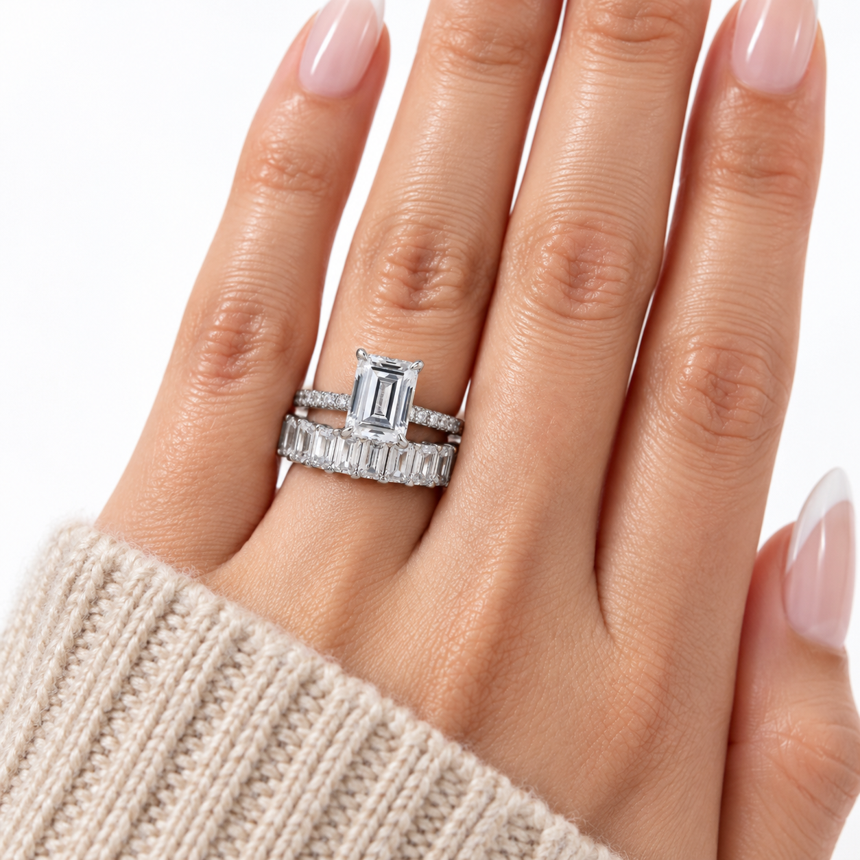A close-up of a hand wearing a silver engagement ring with a rectangular emerald-cut center stone and a matching baguette band, paired with a silver wedding band with small clear stones, set against a plain white background.