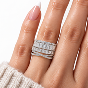 A close-up of a hand wearing a multi-band diamond ring set with baguette and round-cut stones in a silver-toned setting, against a clean white background.