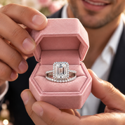 A close-up of a silver engagement ring with a large emerald-cut center stone and a matching halo band, displayed in an open pink velvet ring box held by a person.