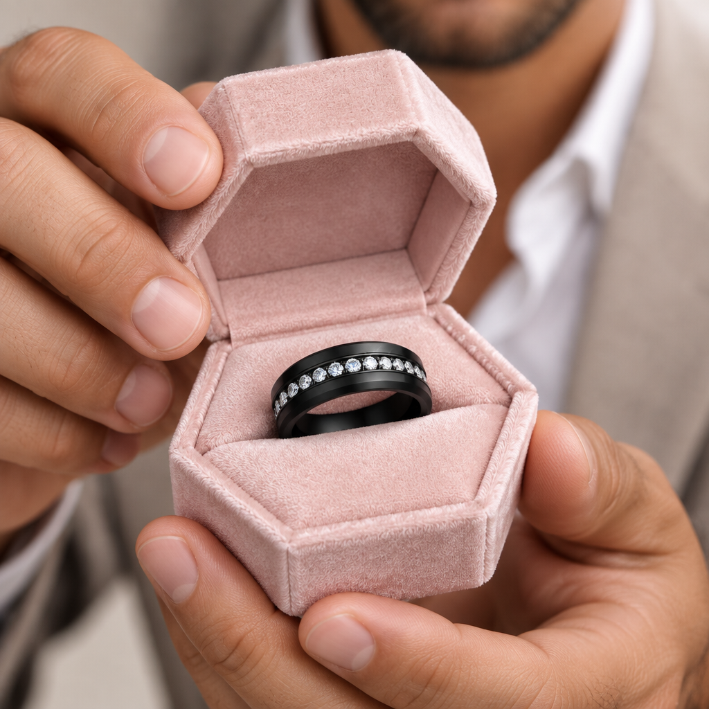 A close-up of a black band ring with a row of clear stones, presented inside an open dusty pink velvet ring box held by a person.