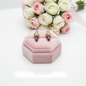 A pair of gold-toned stud earrings with purple crystal points resting on a pink velvet jewelry box, with a blurred bouquet of white and pink roses in the background.