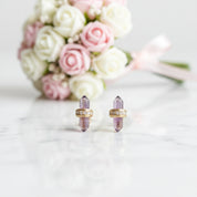 A pair of gold-toned stud earrings with purple amethyst crystal points, displayed on a white marble surface with a blurred bouquet of pink and white roses in the background.