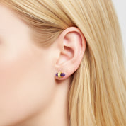 A close-up of a model's ear wearing a small gold hoop earring with a purple and gold crescent-shaped charm.