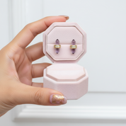 A hand holds an open light pink jewelry box displaying a pair of gold ring settings with small clear stones, against a soft white background.