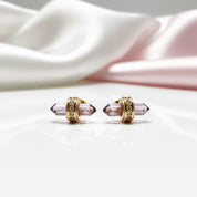 A pair of gold-toned stud earrings with faceted lavender-purple crystal points and small clear stones, displayed on a glossy white surface with soft white and pale pink draped fabric in the background.
