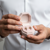 A close-up of a person in a white shirt holding an open light pink jewelry box that displays a pair of gold hoop earrings with purple crystal-like drops.