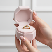 Hand holding a pink jewelry box with a ring inside against a light background