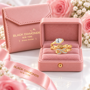Two gold rings displayed in an open pink velvet jewelry box, with a matching pink envelope-style box behind it and pink roses/baby's breath flowers around them. The boxes show the brand name BLACK DIAMONDS NEW YORK.