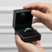 Hand holding a black jewelry box with a green gemstone ring inside against a neutral background