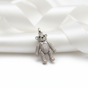 A silver teddy bear charm encrusted with clear rhinestones, resting on a white satin ribbon.