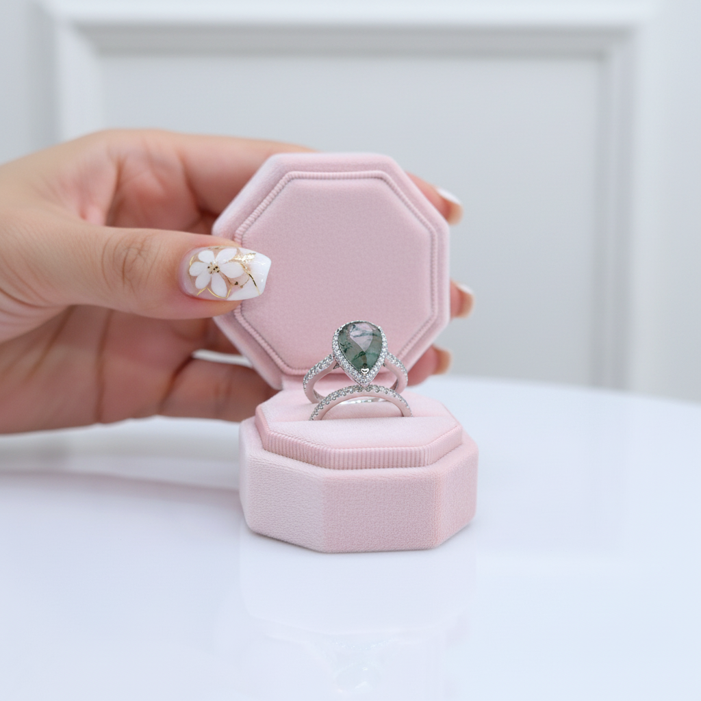 Hand holding a pink jewelry box with a silver ring featuring a green gemstone.