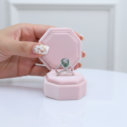 Hand holding a pink jewelry box with a silver ring featuring a green gemstone.