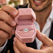 A heart-shaped diamond engagement ring in a silver band, displayed inside an open pink velvet ring box held by a smiling man.