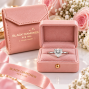 A heart-shaped clear gemstone ring displayed in an open pink velvet jewelry box, with a matching pink branded box and ribbon in the background among soft flowers. Visible brand text on the packaging reads BLACK DIAMONDS NEW YORK.