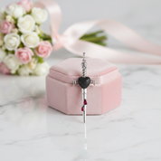 A silver-toned pendant with a black heart and small red teardrop charms, displayed on top of a pink velvet jewelry box on a white marble surface with blurred pink roses in the background.