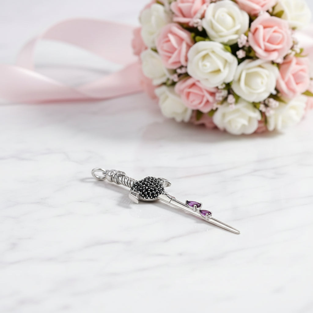 A silver-toned Scottish brooch shaped like a sword, featuring a black crystal-encrusted hilt and two small purple crystals on the blade, displayed on a white marble surface with a pink and white rose bouquet in the background.