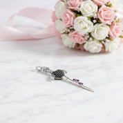 A silver-toned Scottish brooch shaped like a sword, featuring a black crystal-encrusted hilt and two small purple crystals on the blade, displayed on a white marble surface with a pink and white rose bouquet in the background.