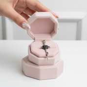 A hand with decorated nails holds an open pink jewelry box containing a silver-toned sword pendant with black and white stones and a small purple accent, set against a clean white background.