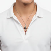 A close-up of a man's tattooed chest wearing a silver sword pendant necklace on a black button-down shirt. The text overlay at the bottom reads 'The chain needs to be purchased separately'.