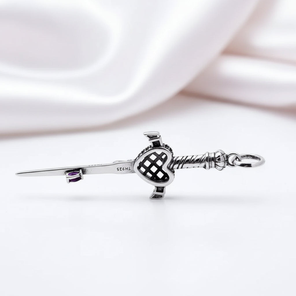 A sterling silver Scottish kiltie pendant shaped like a sword with a heart-shaped guard, resting on a white surface with a soft, draped white satin background.