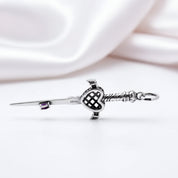 A sterling silver Scottish kiltie pendant shaped like a sword with a heart-shaped guard, resting on a white surface with a soft, draped white satin background.