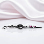 A silver-toned hair claw clip with a textured black ball accent and two small pink gemstones, displayed on a reflective white surface with draped white satin in the background.