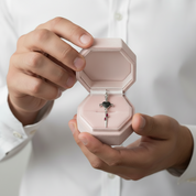 A close-up of a person in a white button-down shirt holding an open light pink jewelry box. Inside the box is a silver-toned pendant with a dark heart-shaped center and small pink gemstone accents.