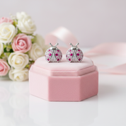 A pair of ladybug-shaped stud earrings with silver-toned metal and pink gemstones, displayed on a pink jewelry box with white and pink roses in the background.