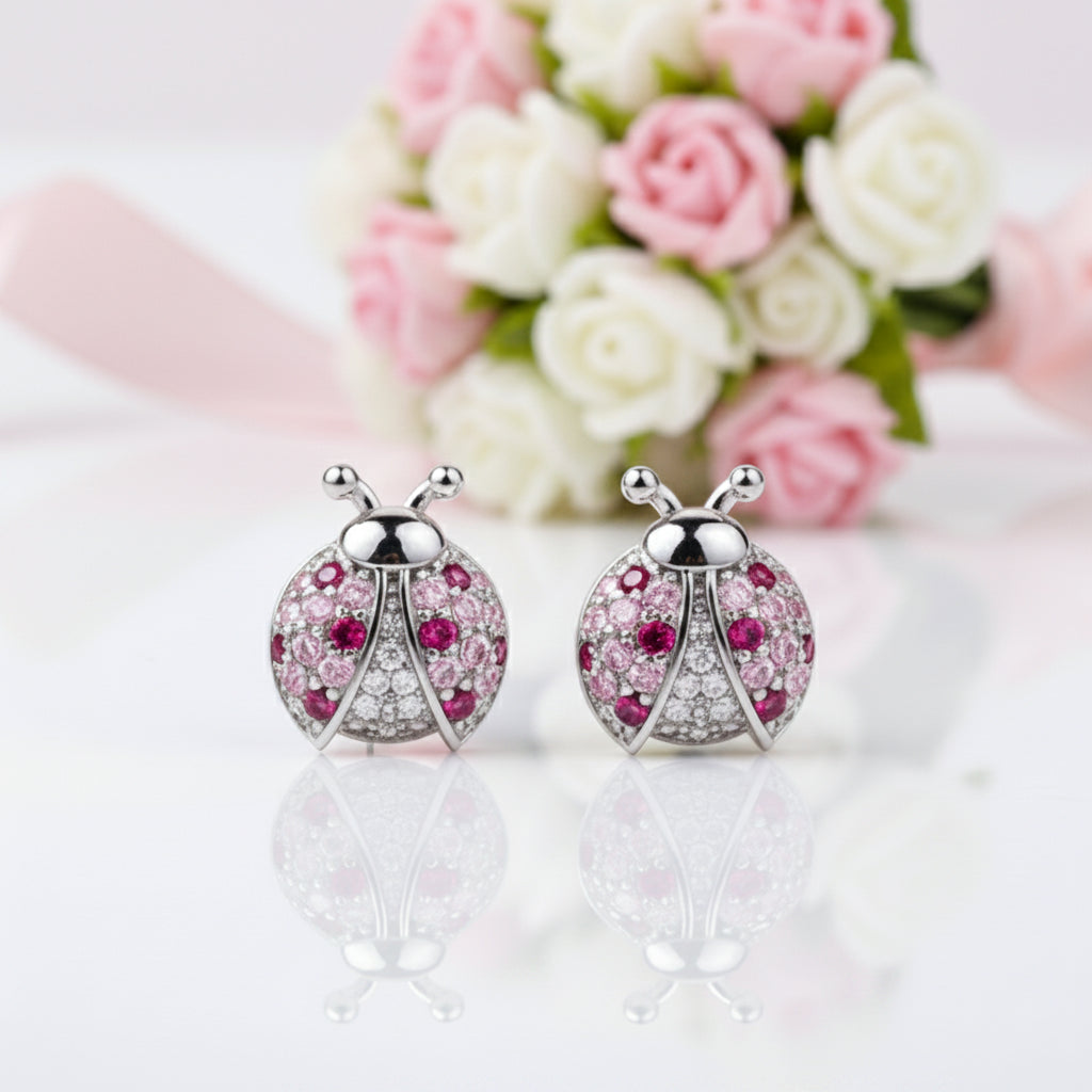 A pair of ladybug-shaped stud earrings with silver-toned metal bodies, pink gemstones, and clear rhinestones, displayed on a reflective white surface with a blurred bouquet of pink and white roses in the background.