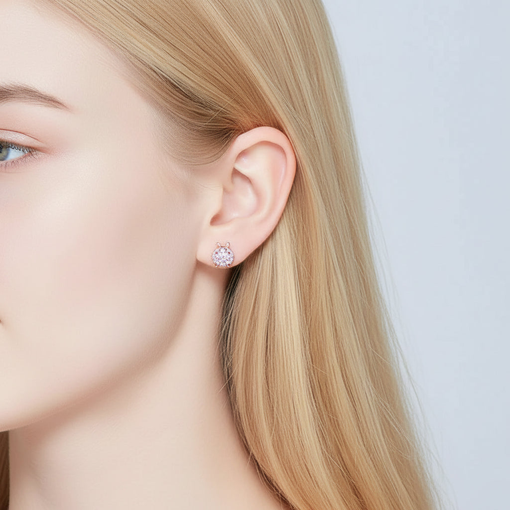 A close-up of a woman's ear wearing a small round stud earring with a silver-toned metal border and a clear central gemstone, set against a soft light gray background.