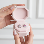 A pair of ladybug stud earrings displayed in an open pink jewelry box, held by two hands with decorated nails against a soft white background.