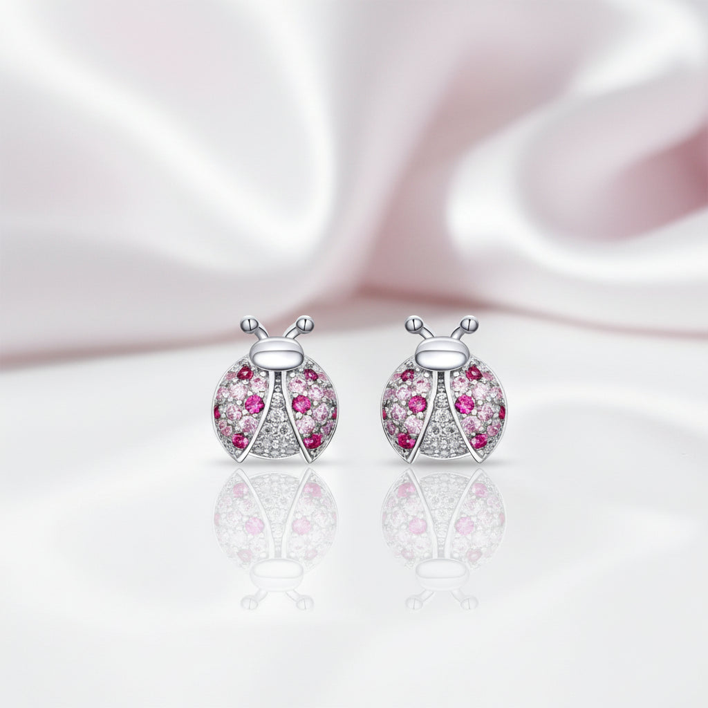 A pair of ladybug stud earrings with silver-toned metal bodies, pink gemstones, and clear pavé accents, displayed on a reflective white surface with a soft pink satin background.