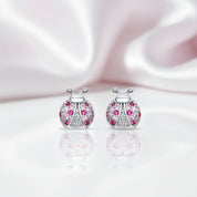 A pair of ladybug stud earrings with silver-toned metal bodies, pink gemstones, and clear pavé accents, displayed on a reflective white surface with a soft pink satin background.