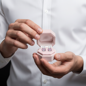 A person in a white dress shirt holds an open light pink jewelry box displaying a pair of small stud earrings with purple and blue stones.