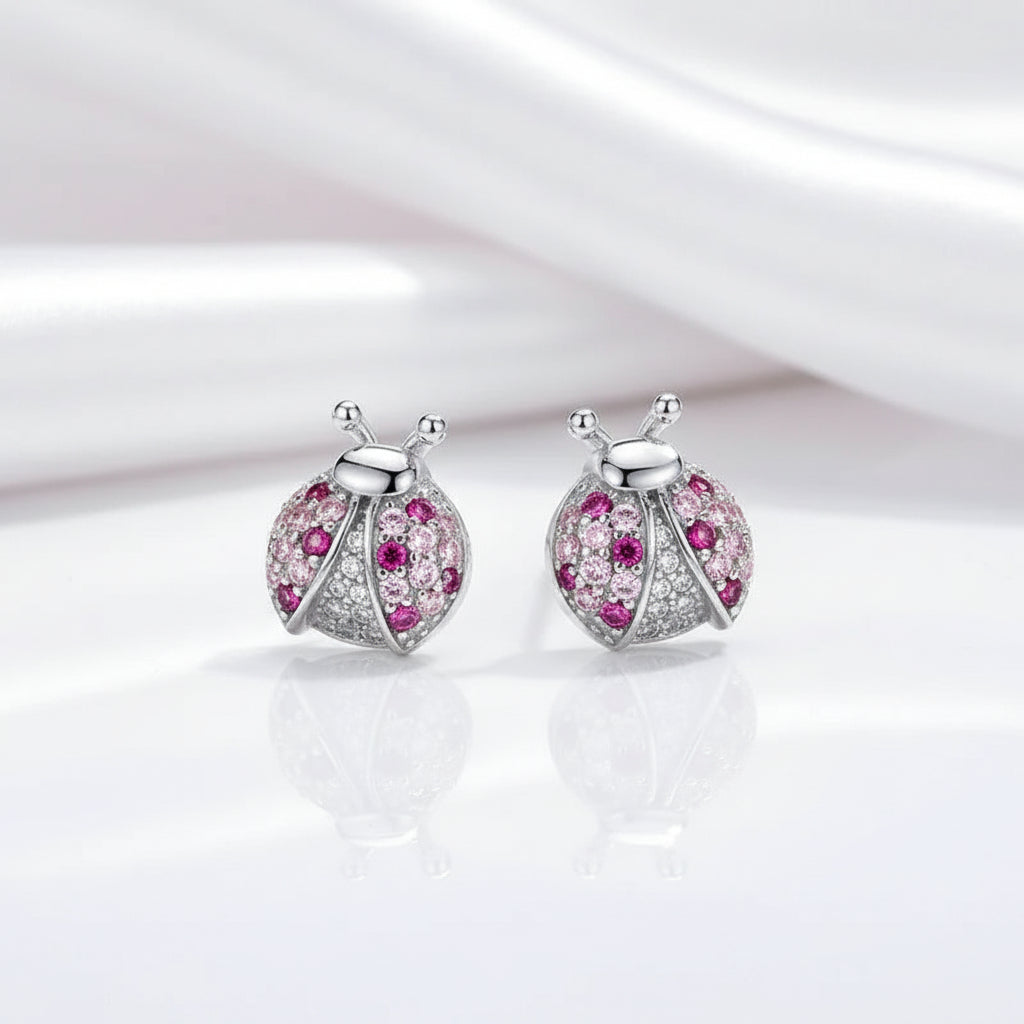 A pair of ladybug-shaped stud earrings in silver-tone metal, decorated with clear pavé stones and pink gemstones, displayed on a reflective white surface with a soft white satin drape in the background.