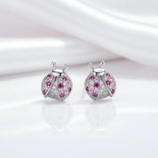 A pair of ladybug-shaped stud earrings in silver-tone metal, decorated with clear pavé stones and pink gemstones, displayed on a reflective white surface with a soft white satin drape in the background.