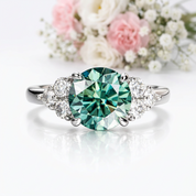 A three-stone diamond ring featuring a large oval-cut emerald-cut green center stone, flanked by two smaller round-cut diamonds on a polished silver-toned band. The ring is displayed on a clean white surface with a soft reflection, and a blurred bouquet of pink and white flowers sits in the background.