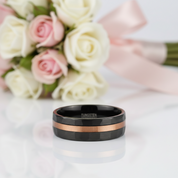 Black tungsten ring with rose gold accents on a white surface with flowers in the background