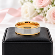 A two-tone metal band ring with a polished gold interior and outer edge, and a brushed silver center band, displayed on a black leather ring box with soft pink and white roses in the background.