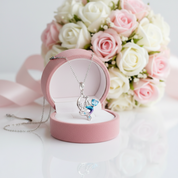 A silver crescent moon and heart pendant necklace with a blue and purple crystal design, displayed in an open pink jewelry box in front of a bouquet of pink and white roses.