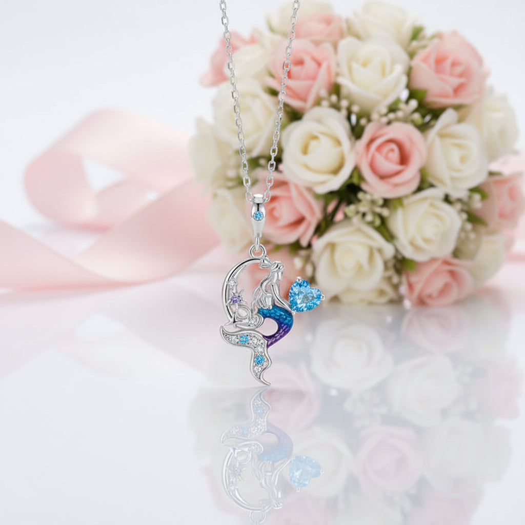 A silver-toned pendant necklace featuring a stylized dancer figure with blue and purple enamel details and small clear stones, displayed against a soft white and blush pink rose bouquet with a pink ribbon.