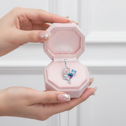 A silver-toned necklace with a blue and white mermaid pendant is displayed inside an open pink octagonal jewelry box, held by two hands with decorated nails against a soft white paneled background.